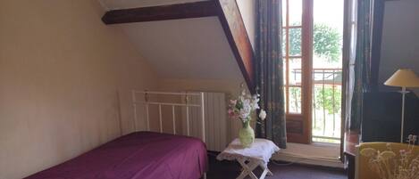 A bedroom with a single bed, a radiator, a window with curtains, a small table with a vase, and a lamp.