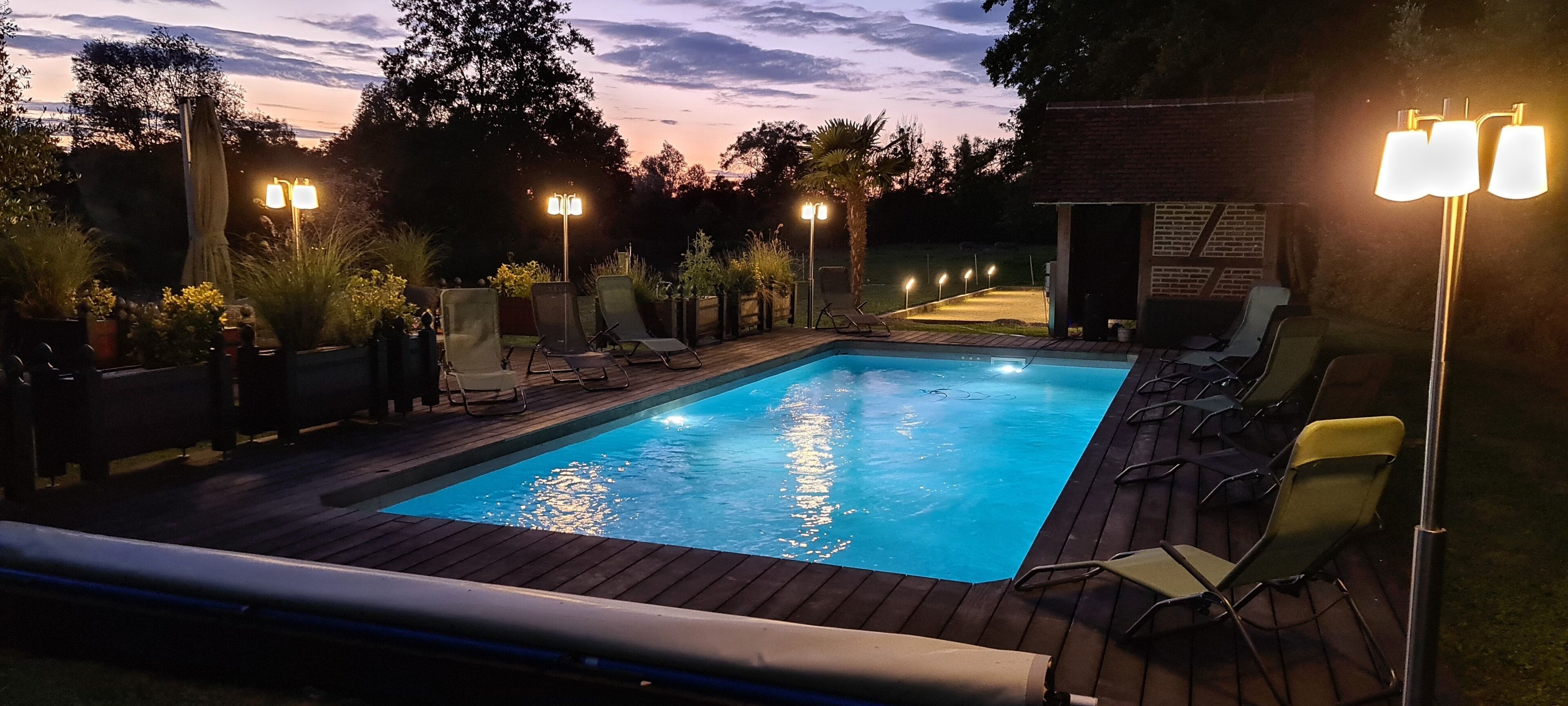 An outdoor swimming pool with lounge chairs and lights, surrounded by plants and a brick structure.