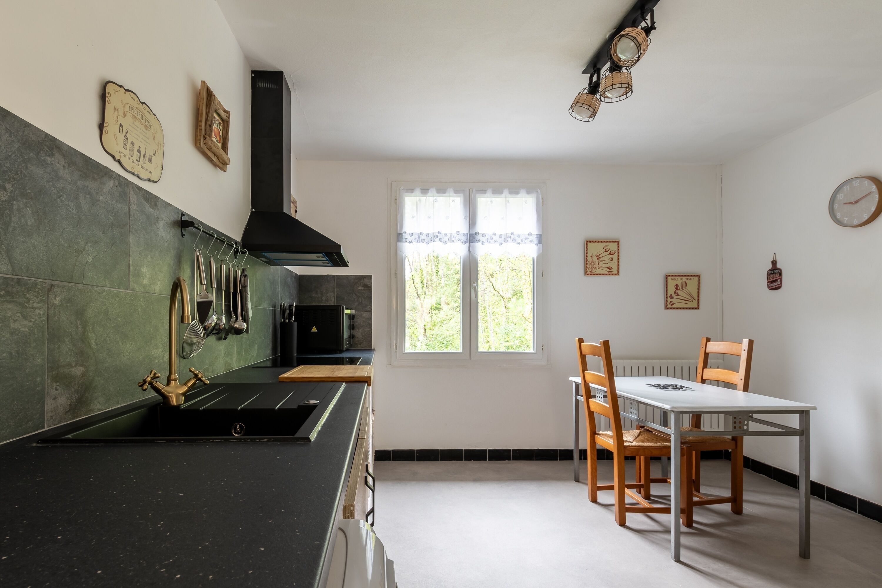 Eine Küche mit schwarzem Arbeitsplatz, einer Edelstahlspüle und einem Fenster mit Blick auf Grünflächen. Ein Esstisch mit Stühlen und eine Uhr sind an der Wand angebracht.