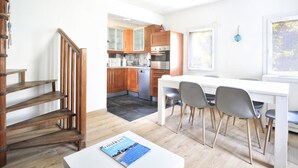 A modern kitchen with wooden cabinets, a dining area with a white table and grey chairs, and a staircase with wooden steps.