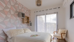A bedroom with a geometric-patterned wall, a bed with a fluffy white cover, a wooden chair, and a large window with a view of the outdoors.