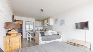 Ein modernes Schlafzimmer mit Bett, Nachttisch und einem kleinen Holztisch. Im Hintergrund ist eine Küchenzeile mit Schränken und einer Arbeitsplatte zu sehen.