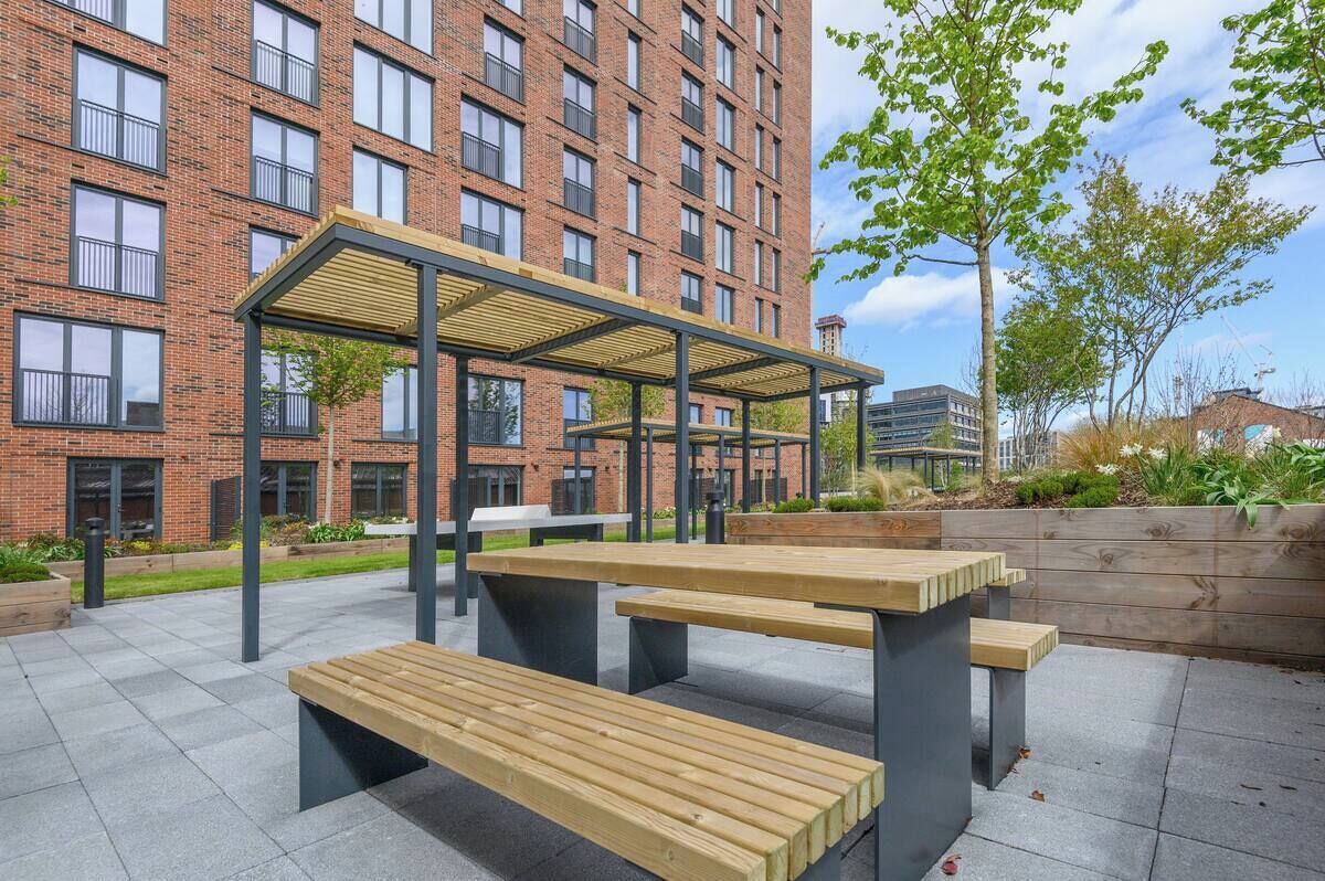 An outdoor seating area with wooden benches and a covered pavilion, set against a brick building with multiple windows.