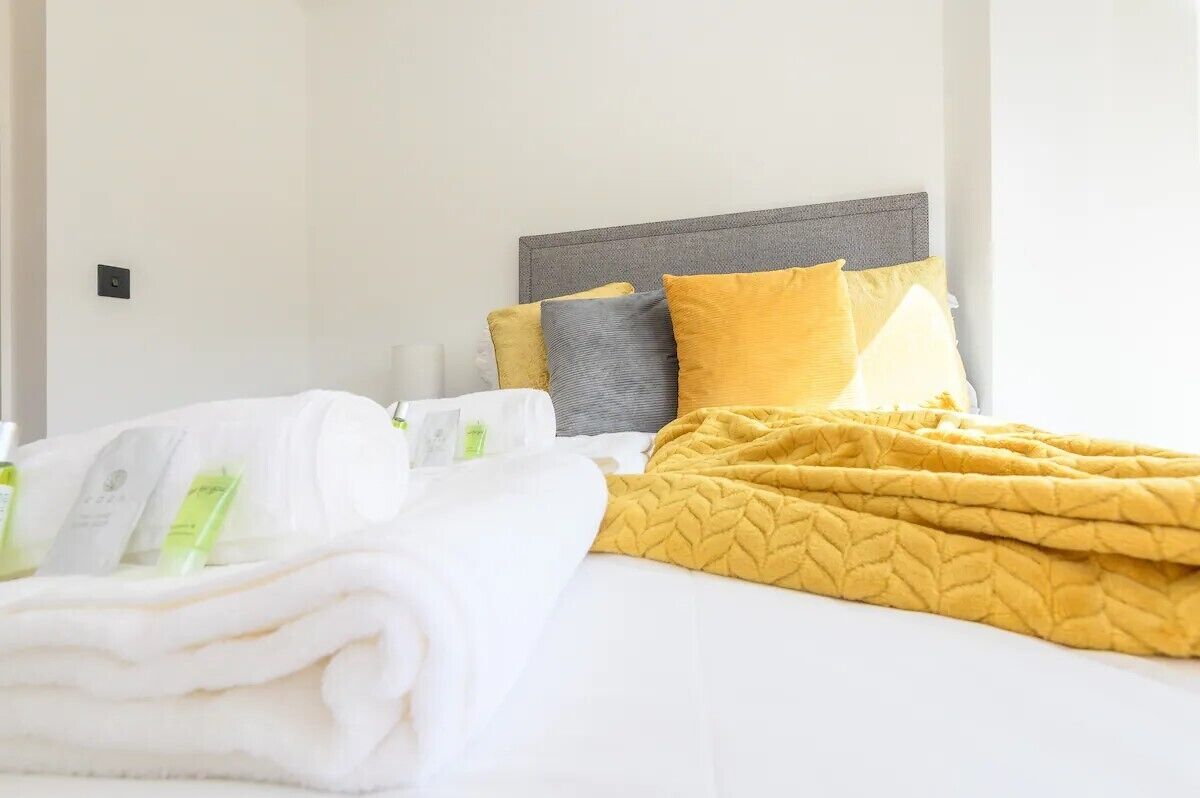 A neatly made bed with a white duvet, a yellow quilt, and two pillows in shades of yellow and gray. There are folded towels and a bottle of lotion on the bed.