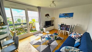 A modern living room with a large window, a blue sofa, a dining table, and a TV.