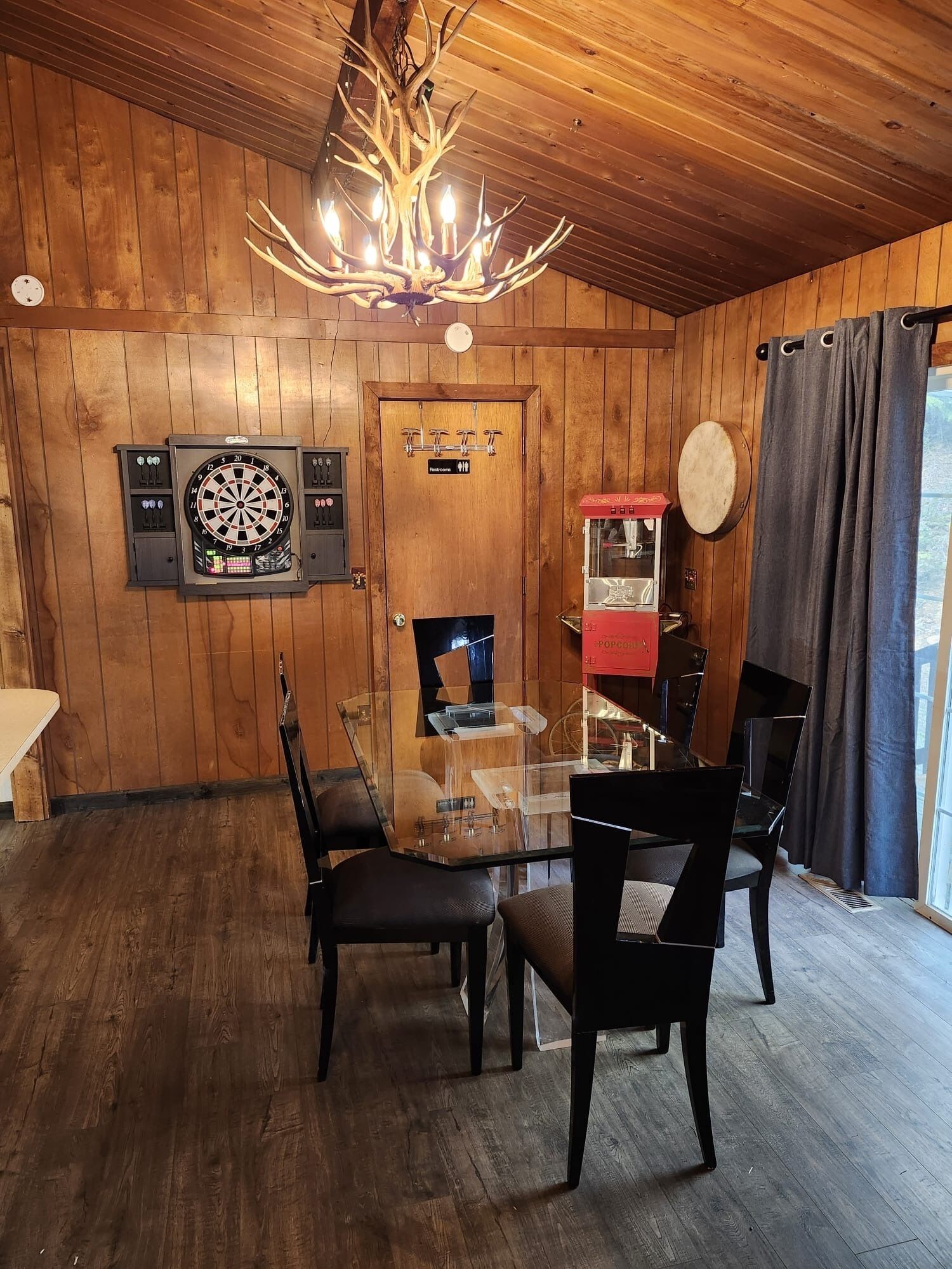 A wooden-paneled room with a glass dining table, chairs, a dartboard, a clock, and a popcorn machine.
