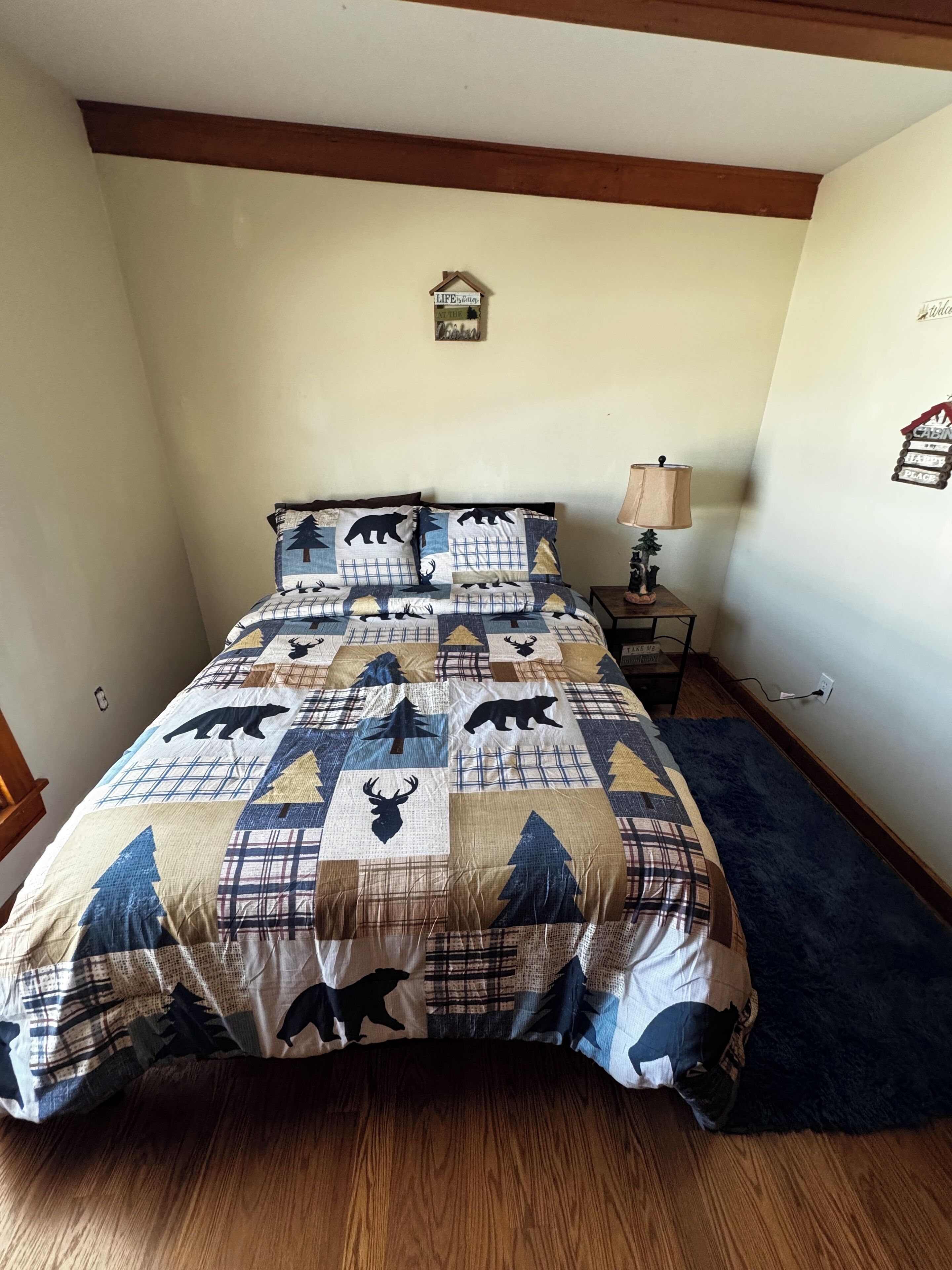 A bedroom with a bed covered in a wildlife-themed quilt, a small bedside table with a lamp, and a wall-mounted decoration.