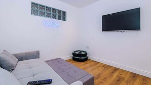 Un salon moderne avec un canapé gris, un téléviseur à écran plat fixé au mur, un néon lumineux affichant le mot « Good Vibes », et une petite table ronde surmontée d’un coussin à motifs.