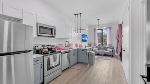Een moderne keuken met roestvrijstalen apparatuur, grijze kastjes en een grijs aanrecht. Er is een zithoek met een grijze bank en een roze stoel.