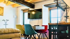 Un salon chaleureux avec un plafond en bois, un canapé, des fauteuils colorés, une table à manger et un téléviseur fixé au mur.