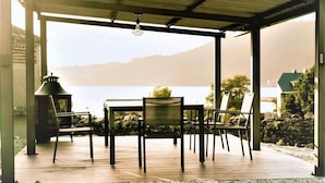Un espace extérieur abrité, aménagé pour les repas, avec une table et des chaises, offrant une vue sur un plan d’eau et des montagnes.