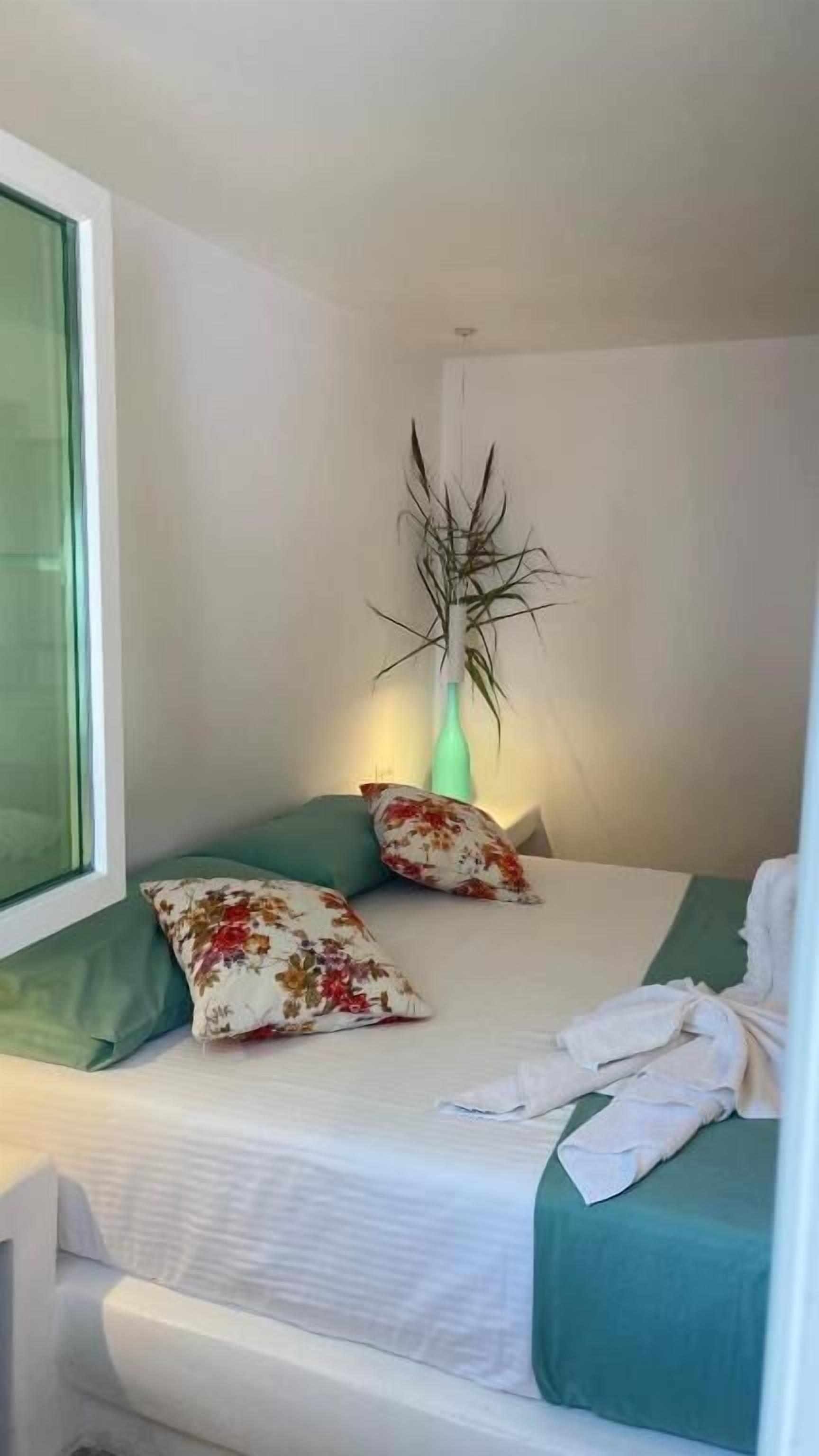 A small, neatly arranged bedroom with a single bed, two pillows, a green bedside table, a window with a view, and a decorative plant.