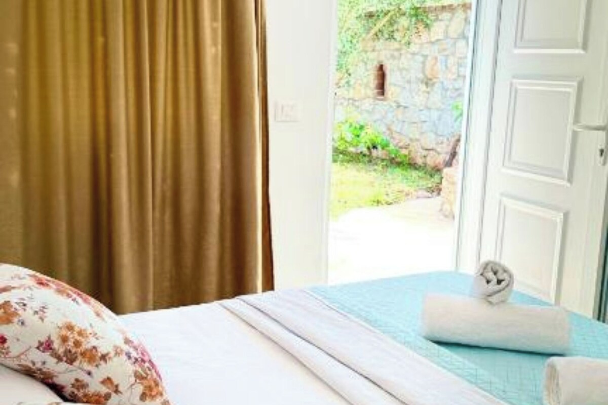 Une chambre à coucher avec un lit, des coussins à motifs floraux, une vue sur un jardin par une porte ouverte et une serviette sur le lit.