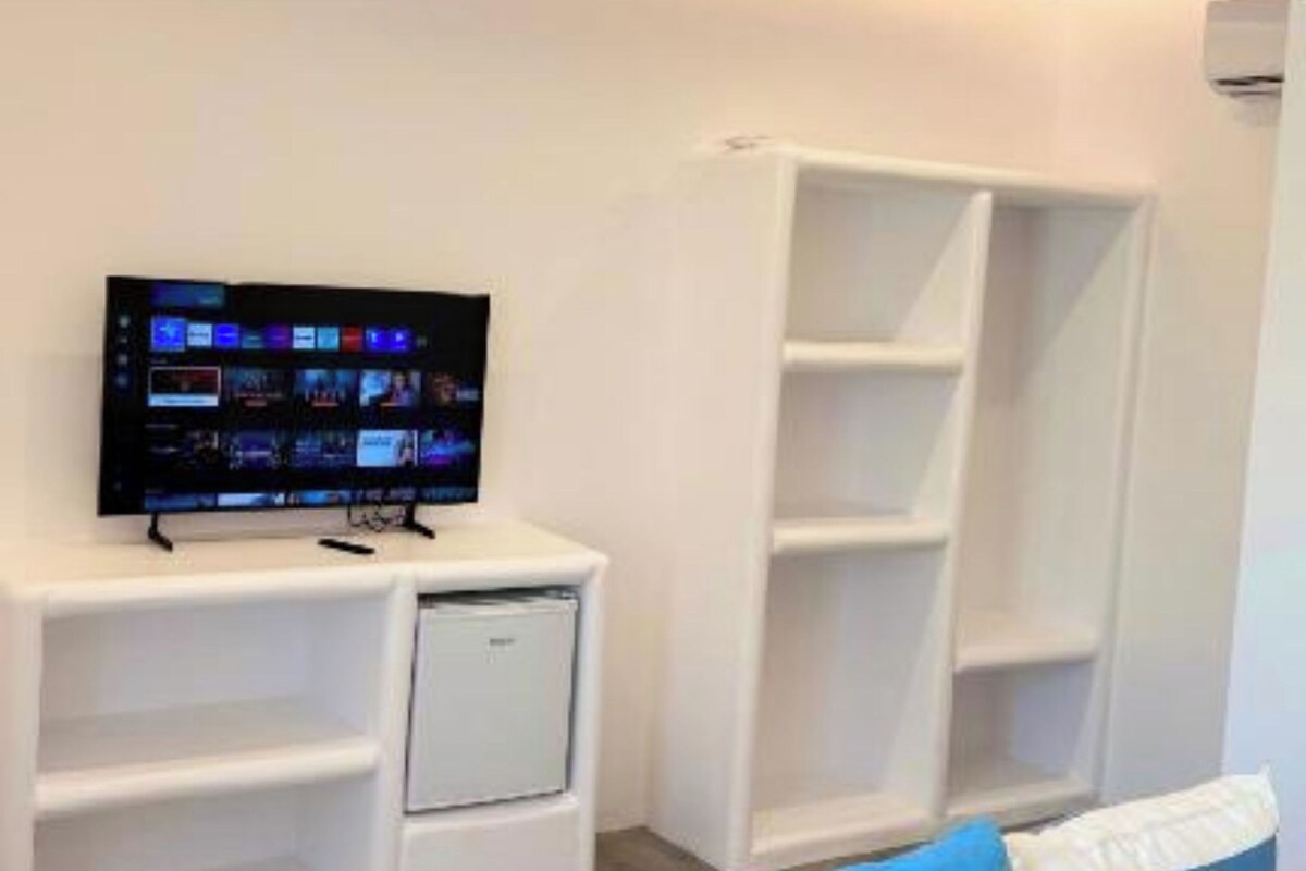 A modern living room with a flat-screen TV, white shelving units, a sofa with patterned cushions, and recessed ceiling lighting.
