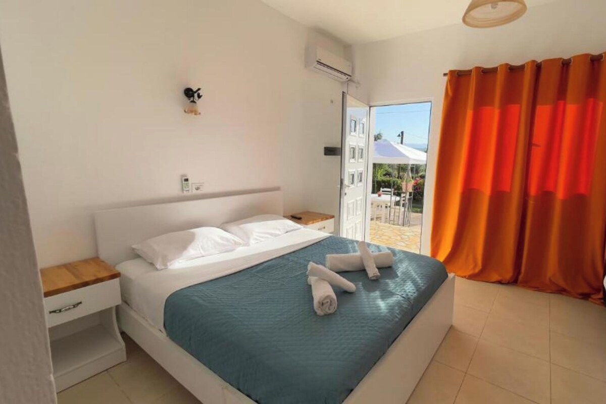 A bedroom with a bed, a nightstand, a drawer, an air conditioning unit, and a view of the outdoors through a door.