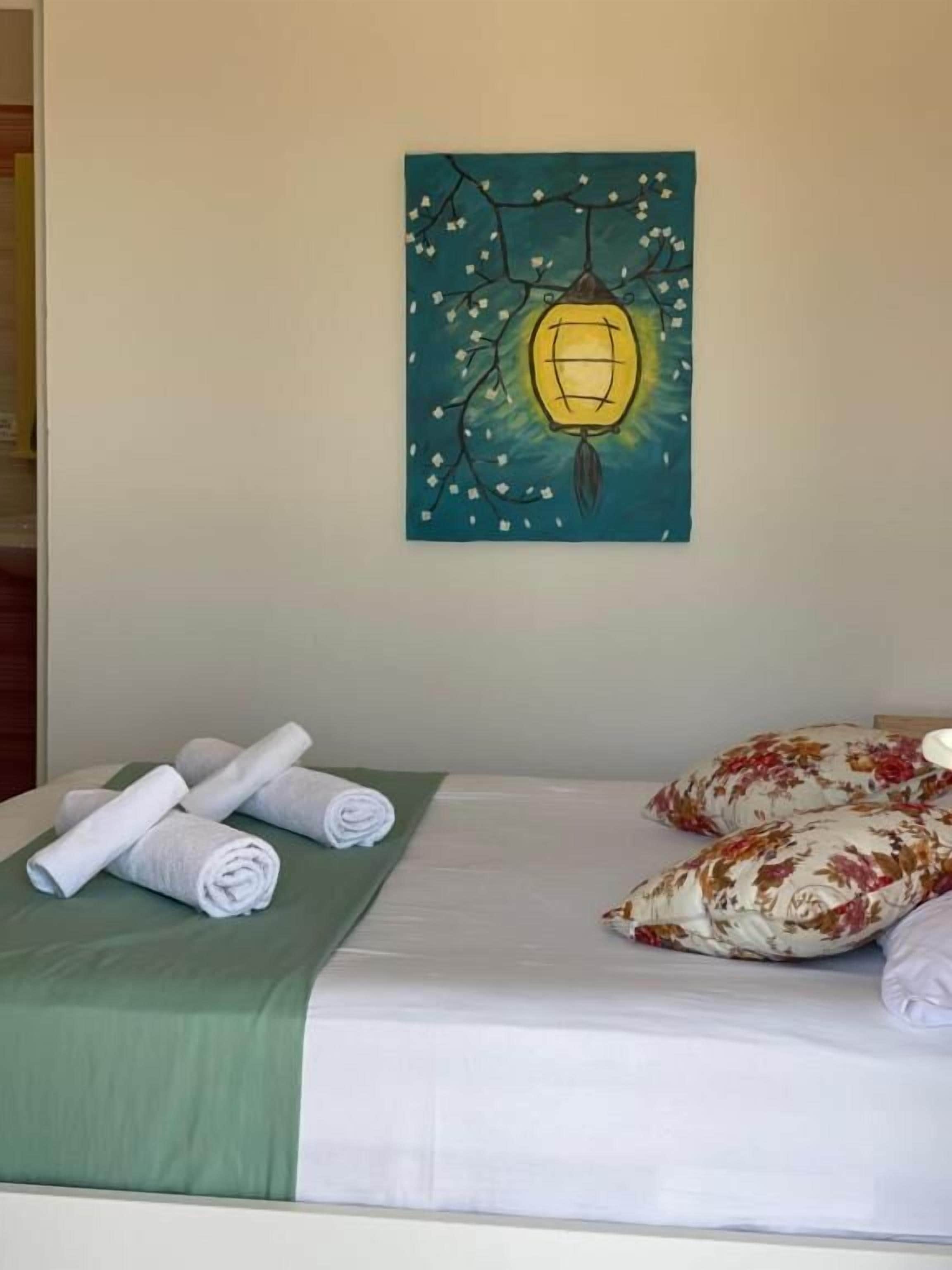 A neatly made bed with rolled white towels and floral pillows, a green bedspread, and a wall-mounted painting of a lantern.