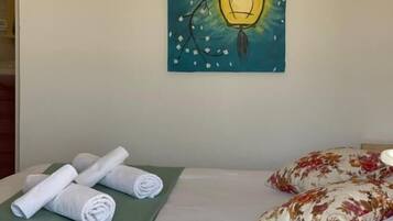 A neatly made bed with rolled white towels and floral pillows, a green bedspread, and a wall-mounted painting of a lantern.