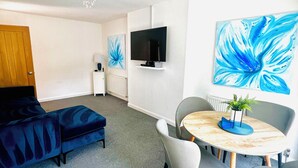 A modern living room with a sofa, a round table with chairs, a TV mounted on the wall, and a large abstract painting. - modern Bungalow with hot tub, garden, bbq & Netflix 10 min drive to city centre (Southampton)