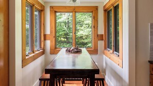 A dining area with a long wooden table and benches, large windows with a view of trees, and a ceiling fan.