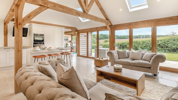 Amplia sala de estar con techo alto y vigas de madera a la vista, cocina moderna y ventanas grandes con vistas al campo.