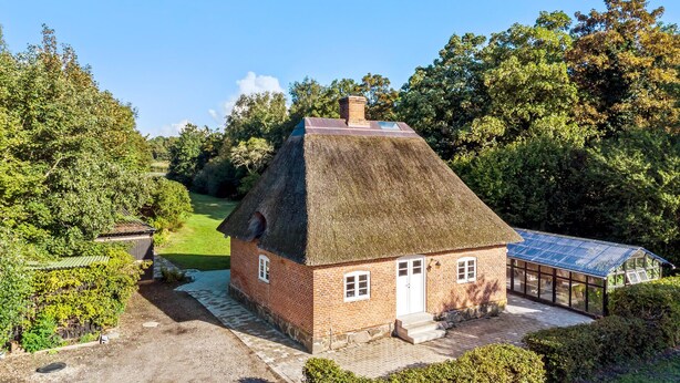 Une maison traditionnelle en briques avec un toit de chaume, un petit abri attenant et une véranda en verre.