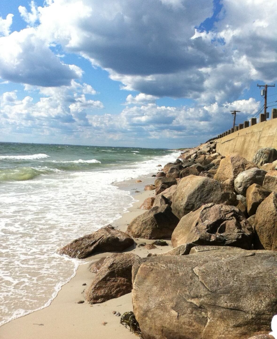 A rocky beach with waves, a concrete wall, and utility poles.