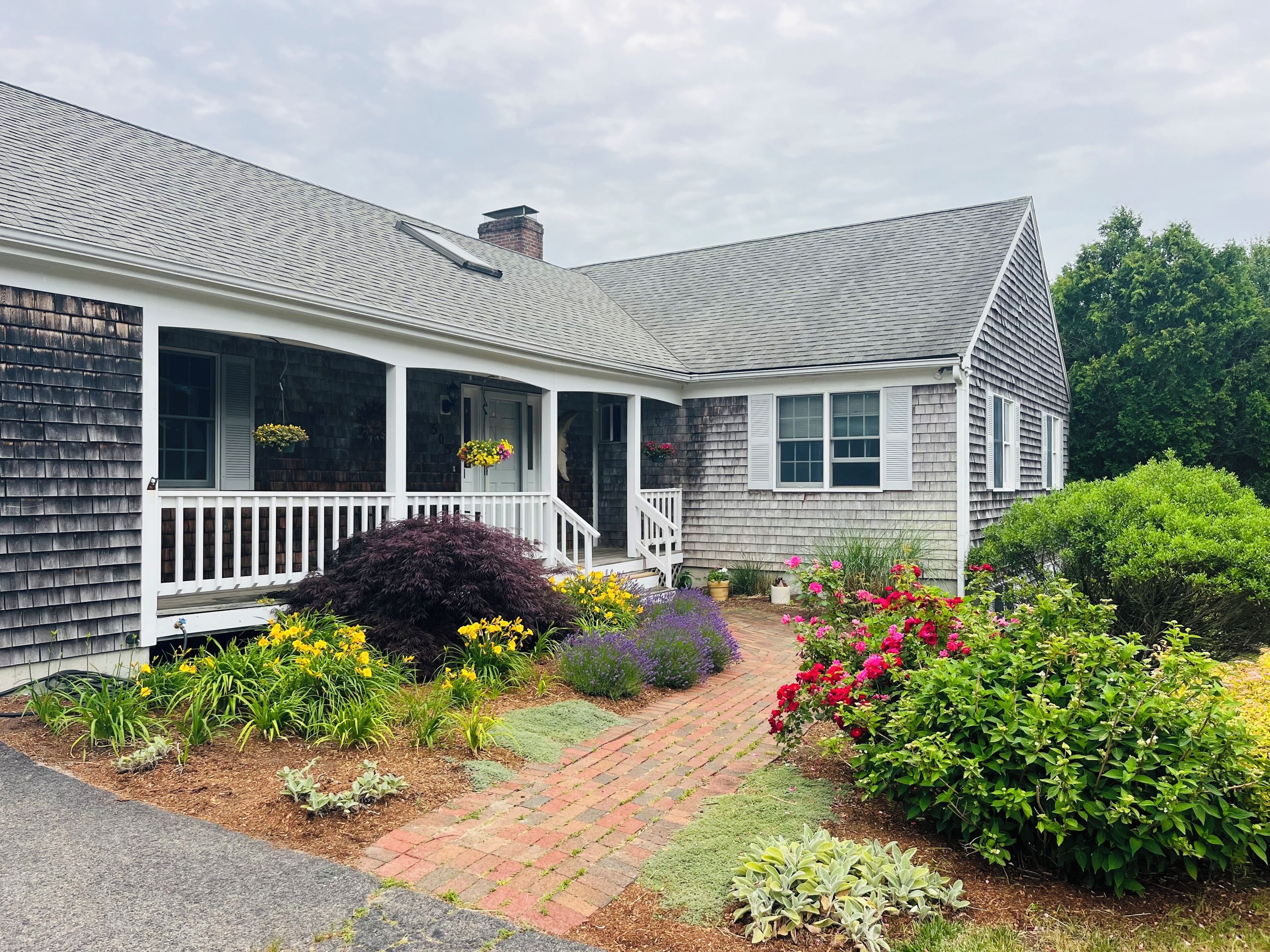 A two-story house with a covered porch, a brick pathway, and various flowering plants.