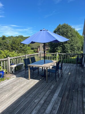 Um deck de madeira com mesa e cadeiras sob um guarda-sol azul, cercado por guarda-corpo de madeira e vegetação.