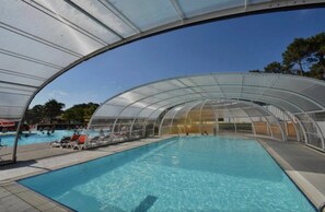 Une piscine couverte avec un toit transparent, entourée de chaises longues et surplombée d’un ciel bleu azur.
