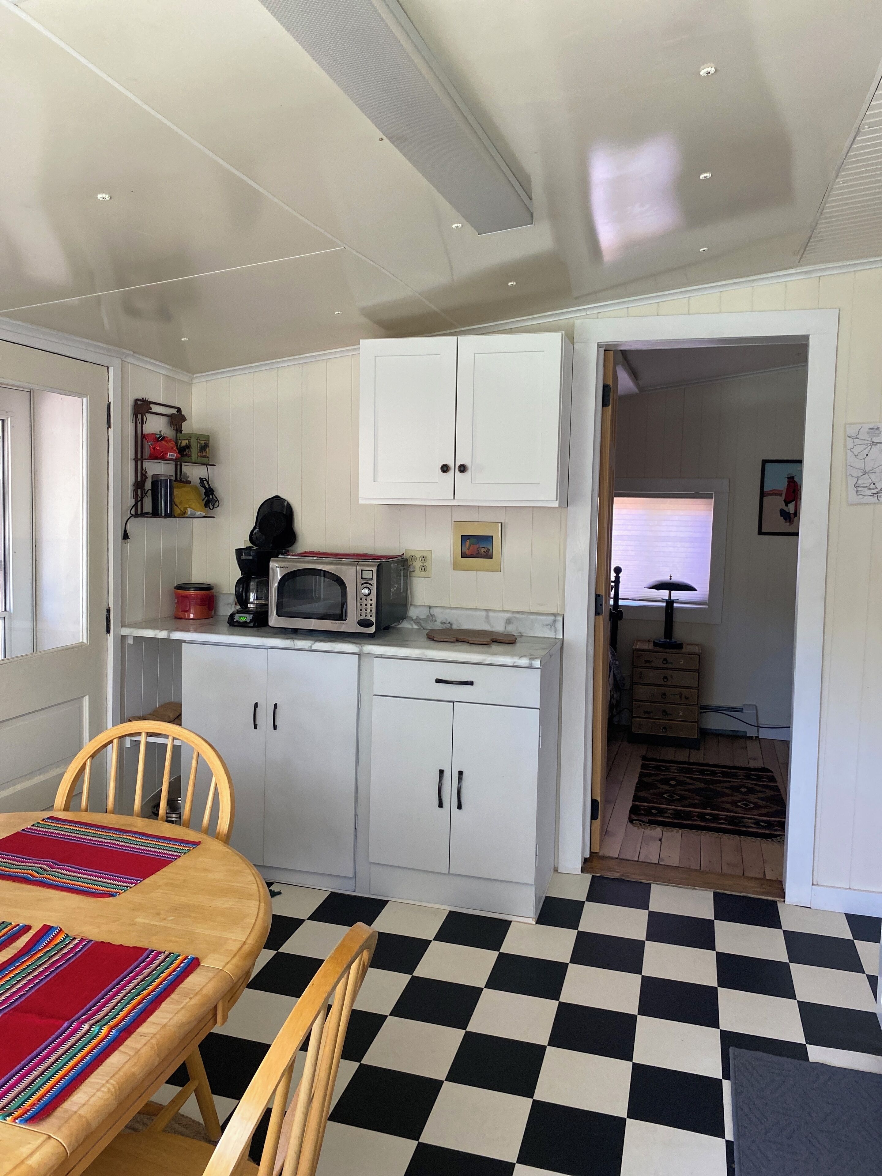 A compact kitchen with white cabinets, a microwave, and a small dining area with a checkered floor and wooden chairs.