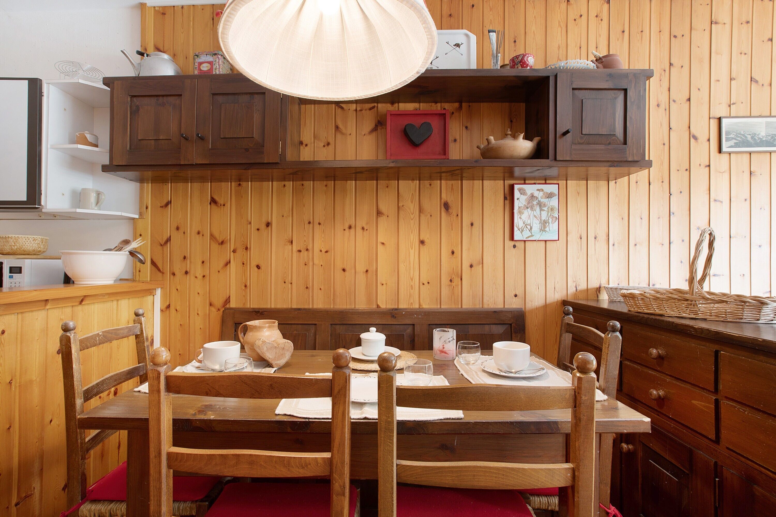 Un comedor de madera con una mesa lista para dos comensales, un banco de madera y un mueble sobre la mesa.