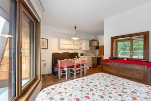 A bedroom with a bed, a wooden dresser, a red table with chairs, a kitchen area, and a window with a view.