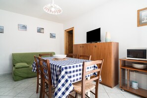 Uma sala de jantar com toalha quadriculada, cadeiras de madeira, um sofá verde, uma televisão e um armário com micro-ondas.