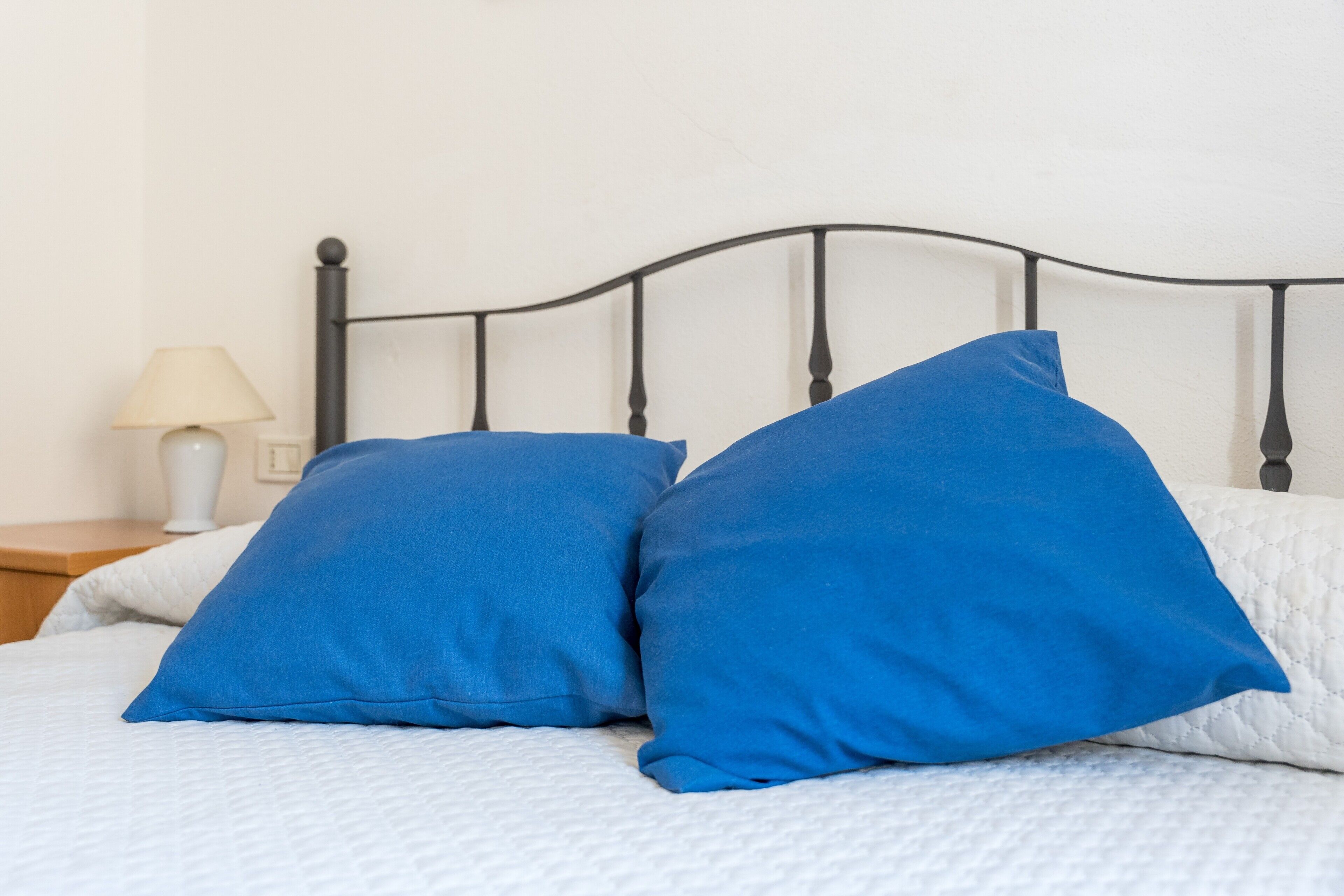 Ein Bett mit weißer Bettdecke, zwei blauen Kissen, einem Nachttisch mit Lampe und einem Metall-Kopfstück.