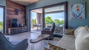Un salon moderne doté d’un téléviseur à écran plat, d’un meuble de rangement en bois, d’un canapé, d’une table basse et d’un balcon offrant une vue sur les arbres et un ciel dégagé.