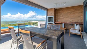 Un espace de restauration extérieur moderne, doté d’une piscine, de mobilier en bois et offrant une vue sur le paysage.