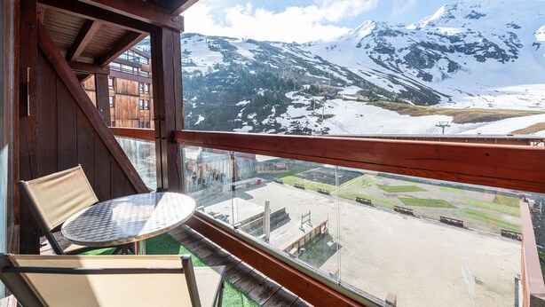 Un balcón con una silla y mesa de madera, que ofrece vistas a una montaña nevada y un estacionamiento.