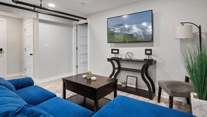A modern living room with a large flat-screen TV mounted on the wall, a dark wooden coffee table, blue sofas, and a grey bench. - Brand NEW Centrally Located with FREE parking and PET friendly!! (Saint Paul)