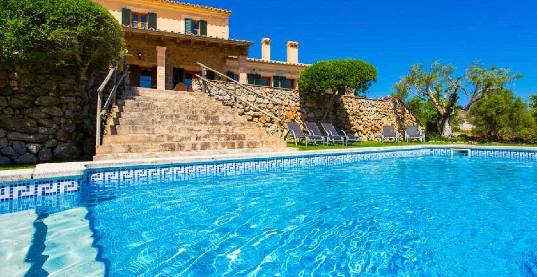 A swimming pool with clear blue water, a stone staircase leading to a building, and a row of lounge chairs.