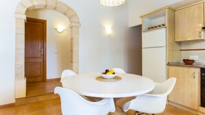 Un comedor con una mesa redonda, sillas blancas, un cuenco de fruta y una cocina con armarios de madera y un refrigerador.