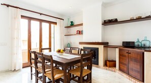 A dining area with a wooden table, chairs, and a bowl of fruit. There is a fireplace, shelves with books and decorative items, and a view of the outside through large windows. - Villa 'Ca Nostra' with Private Pool, Shared Terrace and Wi-Fi (Son Servera)