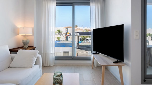A modern living room with a sofa, a TV on a stand, a lamp, and a large window with a view of buildings and a blue tarp. - Apartment 'Apartamento Pachimon' with Shared Pool, Wi-Fi and Air Conditioning (Conil de la Frontera)