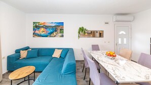 Een moderne woonkamer met een blauwgroene bank, een houten salontafel en een marmeren eettafel met een fruitschaal.