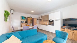 Een modern appartement met een woonkamer, voorzien van een blauwe bank en fauteuils, een eethoek met tafel en stoelen, en een keuken met witte kasten en een televisie.