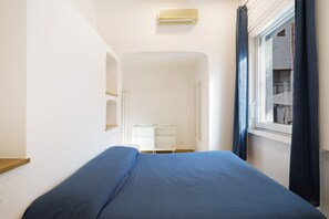Une petite pièce avec un lit, un petit bureau blanc et une fenêtre avec des rideaux.