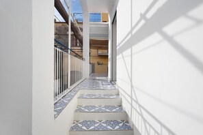 A narrow corridor with patterned tiles, a metal railing, and a view of a building with balconies.