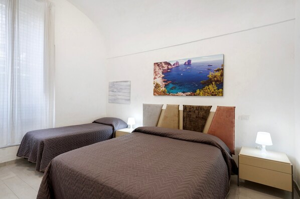 A hotel room with two beds, a painting of a coastal landscape, a nightstand with a lamp, and a window with curtains.
