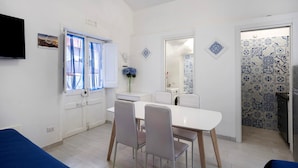 A dining area with a white table and chairs, a window with curtains, a flat-screen TV, and a wall with decorative tiles.