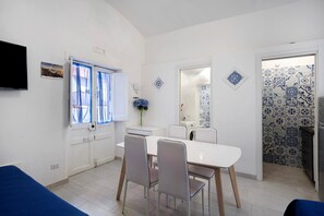 Een eetgedeelte met een witte tafel en stoelen, een raam met gordijnen, een flatscreentelevisie en een muur met decoratieve tegels.