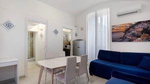 A modern living room with a blue sofa, a dining table with chairs, a kitchen area, and a large window with curtains.